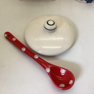 May include: A red ceramic spoon with white polka dots next to a white ceramic lid with a black and white knob. The spoon is angled towards the bottom left, and the lid is in the center of the image.