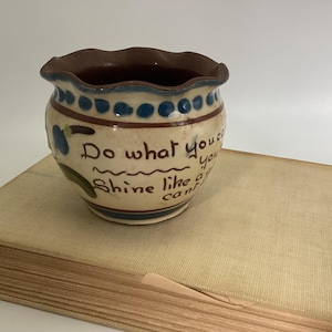 May include: A small, round ceramic pot with a ruffled rim, featuring a cream-colored base with blue and brown accents. The pot has the words "Do what you can, Shine like a can!" written on it. The pot is sitting on top of a book.