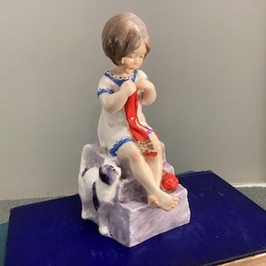 May include: A porcelain figurine of a child with brown hair wearing a white dress with blue trim, sitting on a light purple block and knitting a red scarf. A small black and white cat stands beside the child, and a red ball of yarn sits nearby.