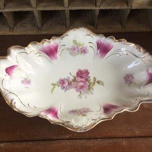 May include: Oval-shaped, white porcelain dish with a scalloped edge, featuring a floral rose design in pink and green. The dish has gold trim and accents of deep pink. A decorative serving piece.
