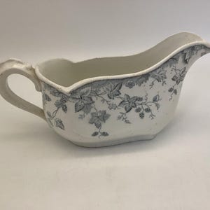 May include: A white porcelain gravy boat with a gray floral design. The boat has a handle and a wide, shallow bowl.