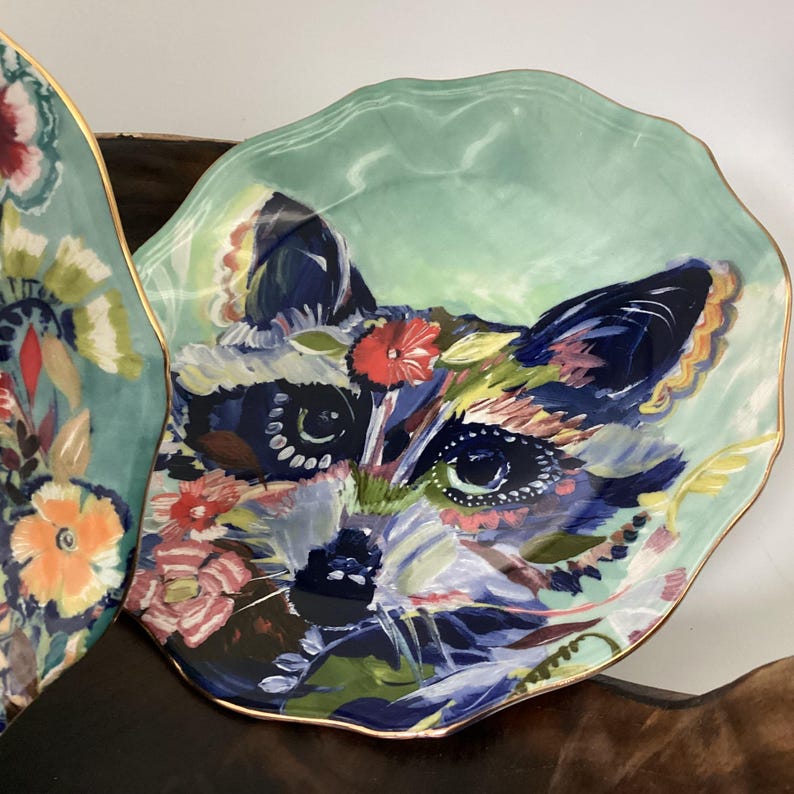 May include: A decorative ceramic plate with a raccoon face design. The plate features a light green background and a gold rim. The raccoon's face is painted with colorful floral accents, including red, pink, and yellow flowers. The plate has a scalloped edge.
