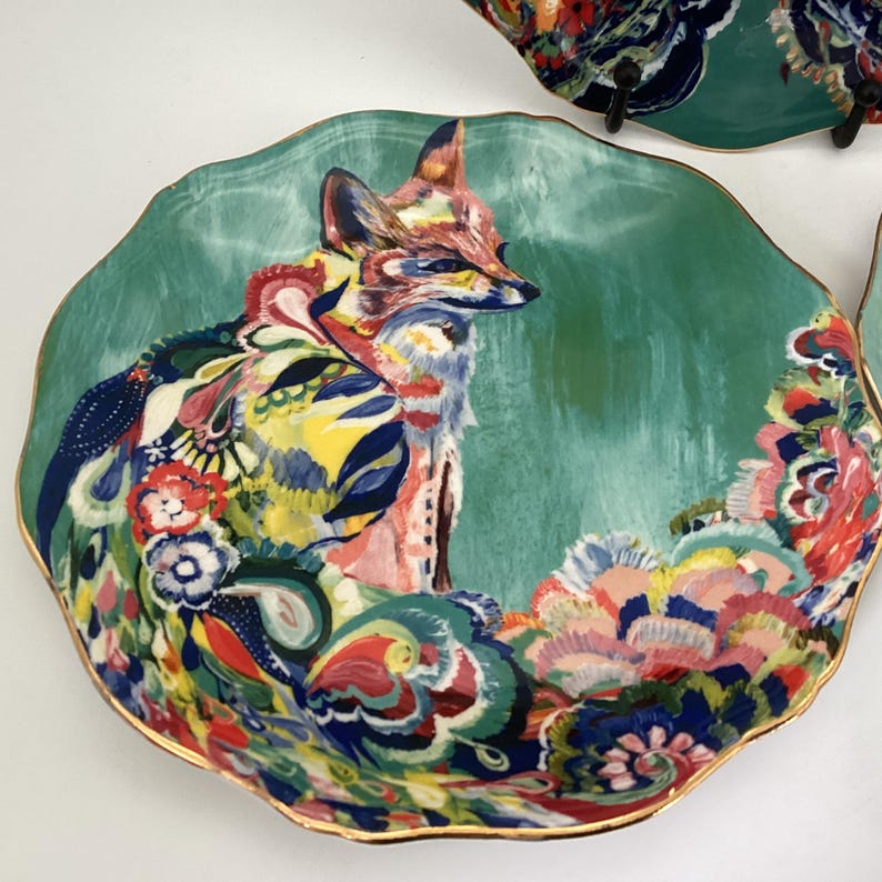May include: Decorative plate with a teal background and a colorful fox illustration. The fox is depicted with vibrant hues of pink, blue, yellow, and red. The plate has a scalloped edge with a gold trim, adding a touch of elegance to the design.