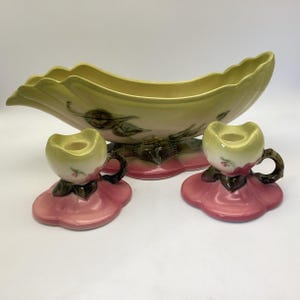 May include: A vintage ceramic centerpiece bowl and two matching candle holders. The bowl is leaf-shaped, with a pale yellow and pink color scheme, and decorative leaf and floral accents. The candle holders are tulip-shaped, with pink bases and yellow tops.