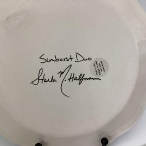 May include: A white ceramic plate with a scalloped edge. The plate features the handwritten text "Sunburst Duo" and the signature "Starla M. Halfmann" in black ink. A small sticker is also visible.