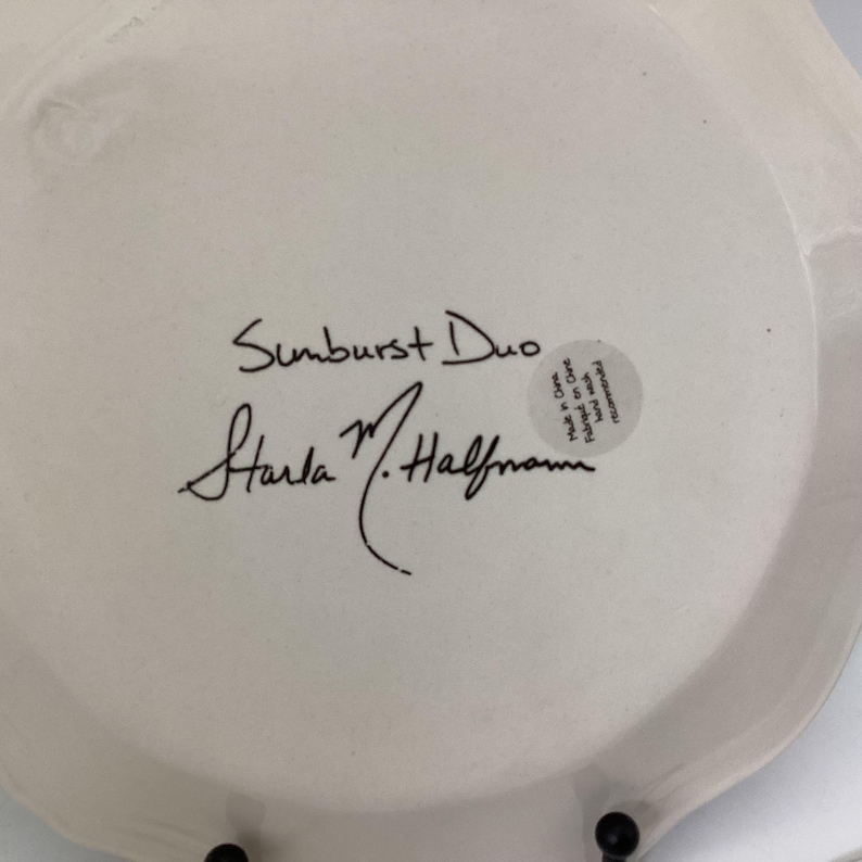 May include: A white ceramic plate with a scalloped edge. The plate features the handwritten text "Sunburst Duo" and the signature "Starla M. Halfmann" in black ink. A small sticker is also visible.