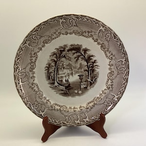 May include: A brown and white decorative plate with a landscape scene in the center. The plate has a detailed border design and is displayed on a wooden stand. The scene depicts trees, buildings, and figures.