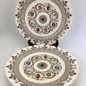 May include: A set of vintage ceramic plates with a cream-colored background and a decorative floral and geometric pattern. The plates feature a scalloped edge and are accented with blue, red, and brown details. The plates are stacked on top of each other.