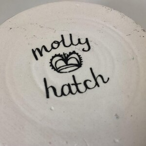 May include: Close-up of the bottom of a white ceramic piece. The black text reads "molly hatch" with a crown symbol between the names. The ceramic has a slightly textured surface.