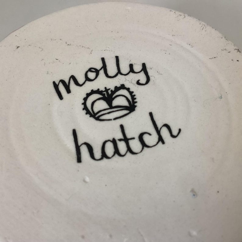 May include: Close-up of the bottom of a white ceramic piece. The black text reads "molly hatch" with a crown symbol between the names. The ceramic has a slightly textured surface.