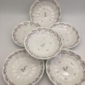 May include: Six white, scalloped-edge saucers. Each saucer features a delicate floral border in blue, green, and red, with a matching floral design in the center. The saucers are arranged in a circular pattern.