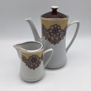 May include: A white ceramic coffee pot and creamer set. The coffee pot has a brown lid and a yellow and brown floral design. The creamer has a matching design and gold trim. The set is on a white background.