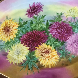 May include: Oval-shaped decorative plate with a hand-painted floral design. The flowers are in shades of yellow, purple, and pink, with green leaves. The plate has a gold rim and a gradient background of pink, yellow, and green.