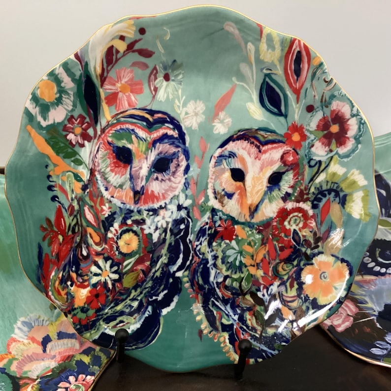 May include: Decorative plate with a teal background and a gold rim. The plate features two colorful owls surrounded by flowers in shades of red, orange, and white. The owls have intricate feather patterns.