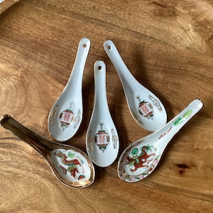 May include: Five white ceramic soup spoons with gold-toned rims. Two spoons feature a red and green dragon design, while three spoons display a red lantern graphic with the Chinese double happiness symbol in black and red ink.