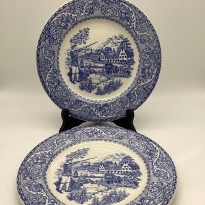 May include: Two blue and white decorative plates. Each plate features a detailed landscape scene with a figure holding a rifle, trees, buildings, and a river. The plates have a decorative border.
