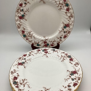 May include: White porcelain plates with a floral design in red, teal, and brown. The plates have a gold rim and a ridged edge. The plates are stacked, with one plate on top, and are likely part of a dinnerware set.