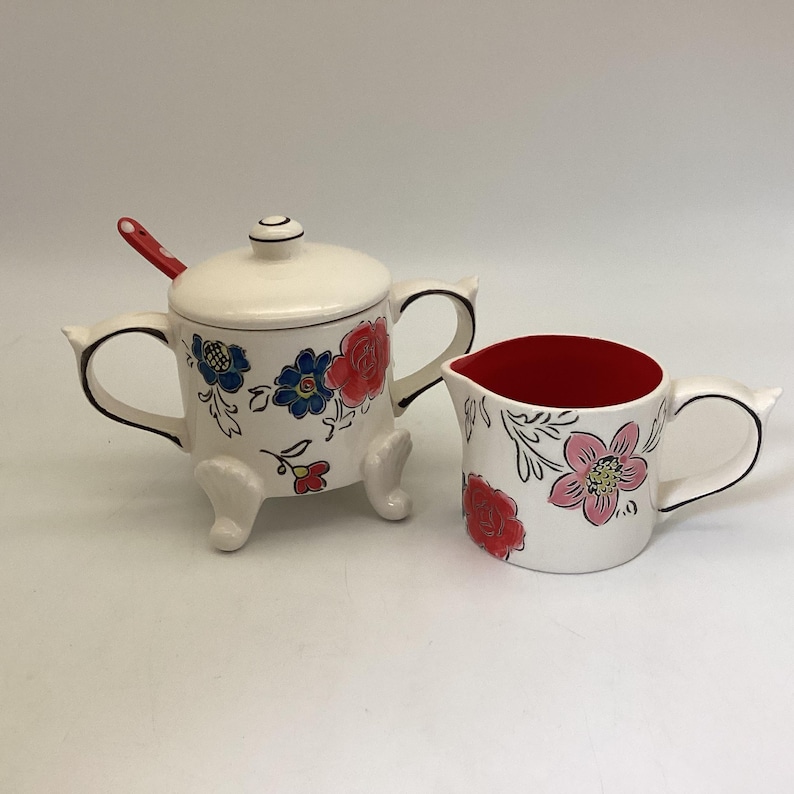 May include: A white ceramic sugar bowl with a lid and a matching creamer. The sugar bowl has a red spoon and is decorated with blue and red floral designs. The creamer has a red interior and floral designs.