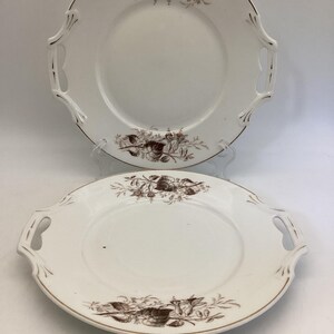 May include: Two white ceramic serving plates with brown floral designs and gold trim. Each plate has two handles. The floral design is at the top and bottom of the plate. The plates are on a white background.