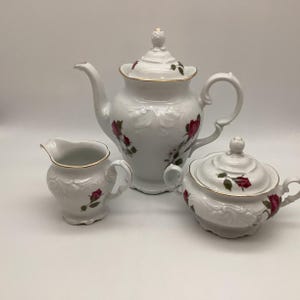 May include: A white porcelain tea set with a teapot, creamer, and sugar bowl. Each piece features a red rose design and gold trim. The set has a classic, elegant aesthetic, perfect for serving tea.