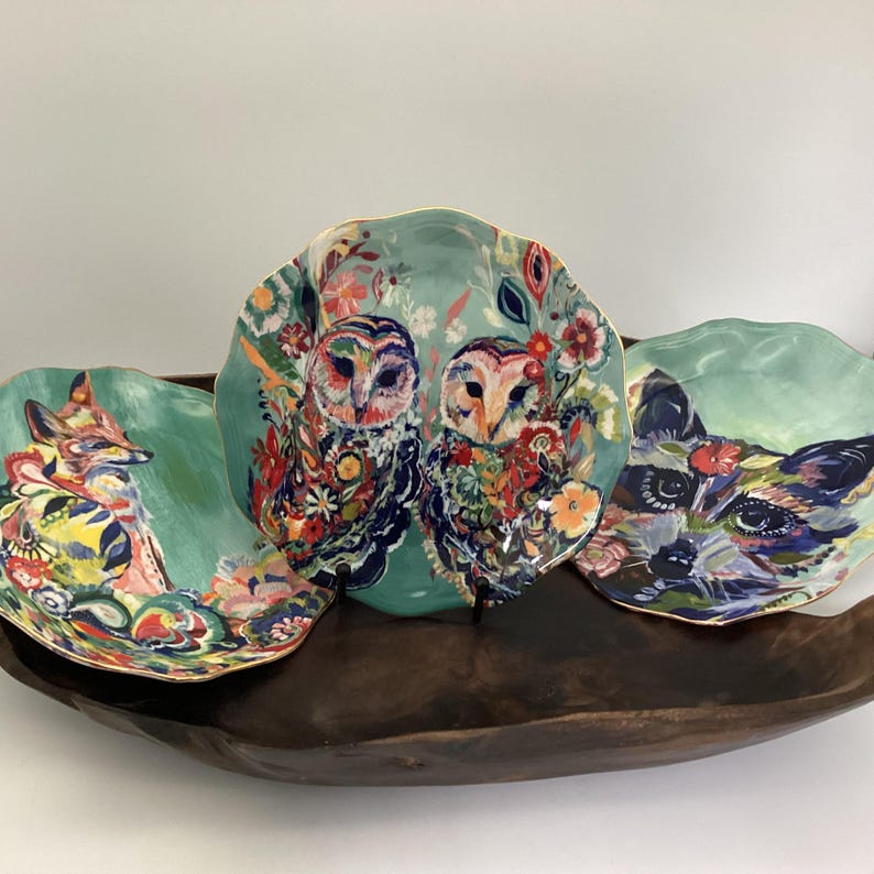 May include: Three decorative ceramic plates with nature-themed designs. One plate features a fox, another two owls, and the third a fox. The plates have a teal background with colorful floral and animal illustrations and gold rims.
