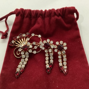 May include: A gold-toned brooch and matching earrings set with red and clear rhinestones. The brooch has a floral design with a central red stone and dangling rhinestone accents. The earrings are clip-on style with dangling rhinestone chains.