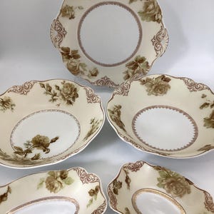 May include: A set of five ceramic dishes featuring a cream-colored base with brown rose floral patterns and ornate scrollwork borders. The collection includes one large round serving plate, two medium round bowls, and two smaller oval-shaped dishes.