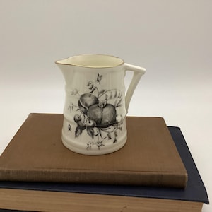 May include: A small, white ceramic pitcher with a handle and a gold rim. The pitcher is decorated with a black and white illustration of apples and cherries. The pitcher is sitting on top of two books.