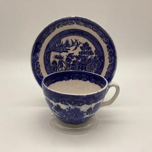May include: A blue and white Willow pattern teacup and saucer set. The cup and saucer feature a detailed landscape design with a blue and white color scheme. The cup has a handle and sits on a clear glass base.