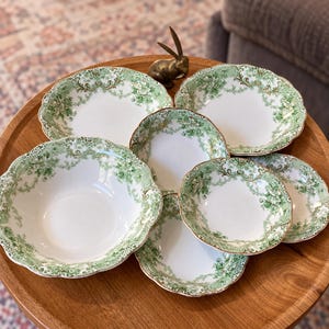 May include: A set of six white ceramic bowls featuring a scalloped edge and a green floral transferware pattern with intricate scrollwork and leaf details. The collection includes one large bowl and five smaller bowls arranged on a wooden surface.