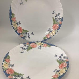 May include: Two white oval plates with a blue scalloped edge. The plates are decorated with a floral pattern featuring pink, yellow, and blue flowers and green leaves. The plates are likely for serving.