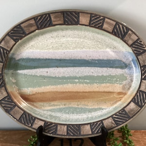 May include: An oval ceramic platter featuring a central glaze pattern of horizontal bands in shades of white, teal, light green, and tan. The wide rim is decorated with a repeating geometric pattern of dark brown squares with diagonal lines.