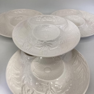 May include: A set of four white ceramic serving dishes. Each dish features a central dip bowl surrounded by a larger, circular serving area. The dishes have a raised floral and leaf pattern, adding texture and detail to the design.