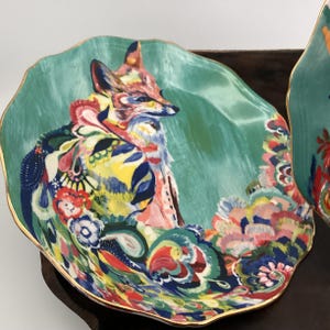 May include: A decorative plate with a teal background and a colorful fox illustration. The fox is depicted with vibrant hues of pink, yellow, and blue. The plate has a gold rim and a unique, wavy shape.