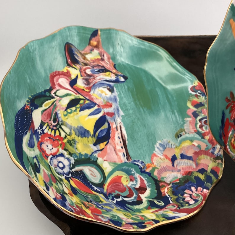 May include: A decorative plate with a teal background and a colorful fox illustration. The fox is depicted with vibrant hues of pink, yellow, and blue. The plate has a gold rim and a unique, wavy shape.
