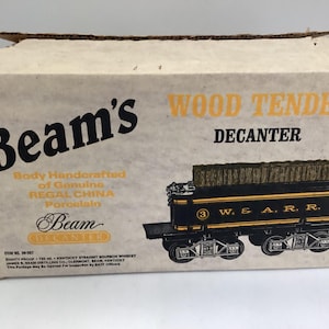 May include: A vintage Beam's Wood Tender Decanter box. The box features the Beam's logo and text, along with an illustration of a black and gold train car. The box is made of cardboard.