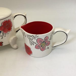 May include: White ceramic creamer with a red interior and floral design. The creamer features a pink flower with a yellow center and red roses. The handle is white with a black outline. A matching sugar bowl is partially visible.