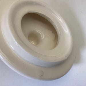 May include: Off-white ceramic lid with a recessed, oval-shaped interior. The lid has a rounded, raised edge and a central, circular depression. The surface appears smooth with subtle variations in color.