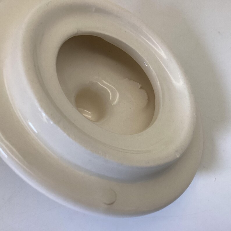 May include: Off-white ceramic lid with a recessed, oval-shaped interior. The lid has a rounded, raised edge and a central, circular depression. The surface appears smooth with subtle variations in color.