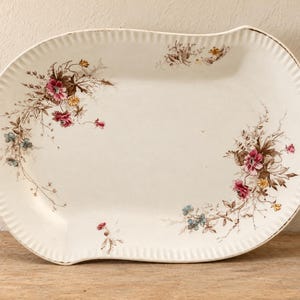 May include: An oval ceramic serving platter with a cream-colored base and a fluted, scalloped edge. The platter features delicate floral illustrations in shades of pink, blue, and brown, arranged in clusters along the left and right sides.
