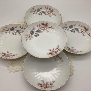 May include: Five white porcelain plates with a scalloped edge and gold trim. Each plate features a floral design with blue and pink flowers and brown stems. The plates are arranged on a white lace doily.