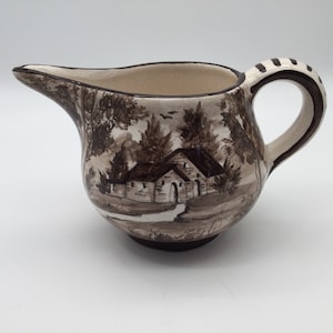 May include: A brown and white ceramic creamer with a hand-painted scene of a house and trees. The creamer has a rounded body and a handle.