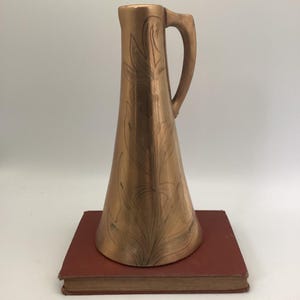 May include: A copper-colored pitcher with a handle and a flared base, adorned with etched floral designs. The pitcher is set on a red book. The pitcher is approximately 10 inches tall.