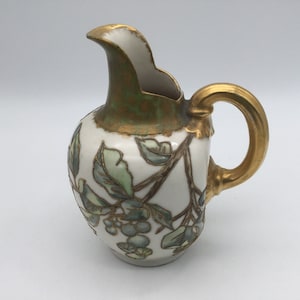 May include: A small white ceramic pitcher with a gold handle and a green and gold floral design. The pitcher features a floral pattern with green leaves and gray berries.