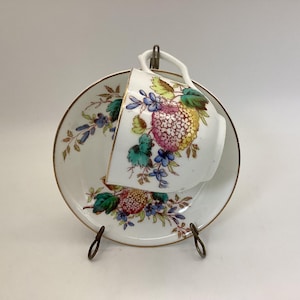 May include: A white porcelain teacup and saucer with a floral design in shades of pink, yellow, green, and blue. The cup and saucer have a gold trim around the edge.