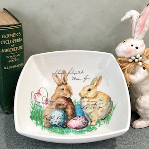 May include: A white square ceramic bowl featuring a printed illustration of two rabbits, one brown and one light tan, sitting in green grass with pink flowers and colorful speckled eggs. The bowl includes cursive script text above the rabbits.