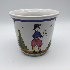 May include: White ceramic planter with a blue rim and a hand-painted design of a person in a blue shirt and red pants holding a stick. The design also includes green trees and brown accents.