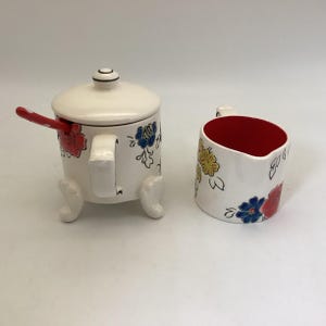 May include: A white ceramic sugar bowl with a lid and a matching creamer. Both pieces are decorated with colorful floral designs in red, blue, and yellow. The sugar bowl has a red and white polka dot spoon.