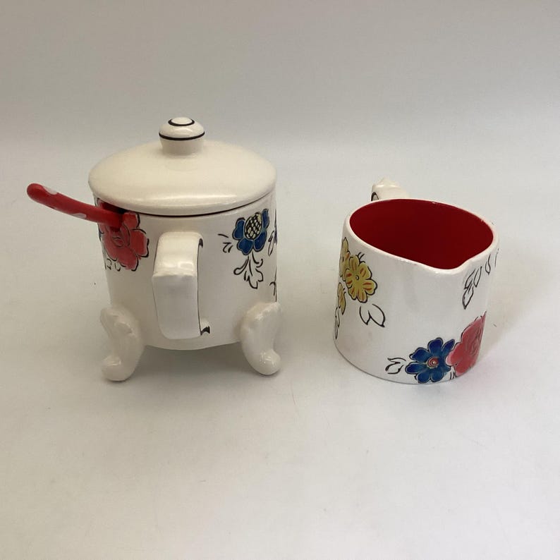 May include: A white ceramic sugar bowl with a lid and a matching creamer. Both pieces are decorated with colorful floral designs in red, blue, and yellow. The sugar bowl has a red and white polka dot spoon.