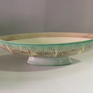 May include: A ceramic serving bowl with a boat-like shape. The bowl has a light green rim and a white base. The exterior features a brown and beige pattern. The interior is beige.